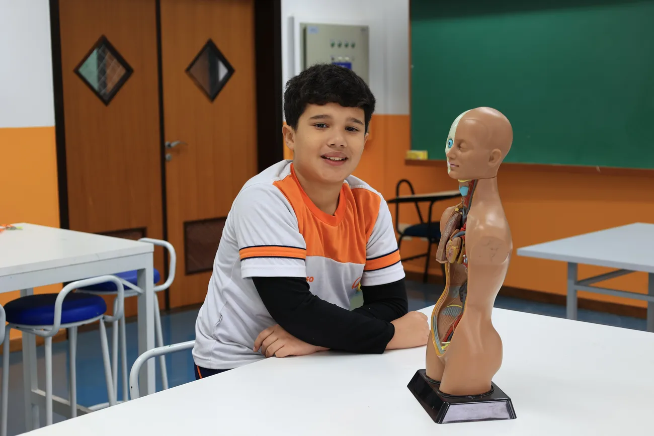 Aluno do Ensino Fundamental do Colégio Acesso sorrindo ao lado de modelo anatômico em sala de aula